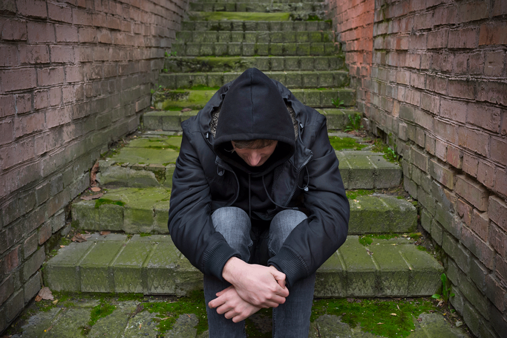 young man in hoodie with head bowed seated outside on concrete steps wondering what are 5 facts about heroin addiction
