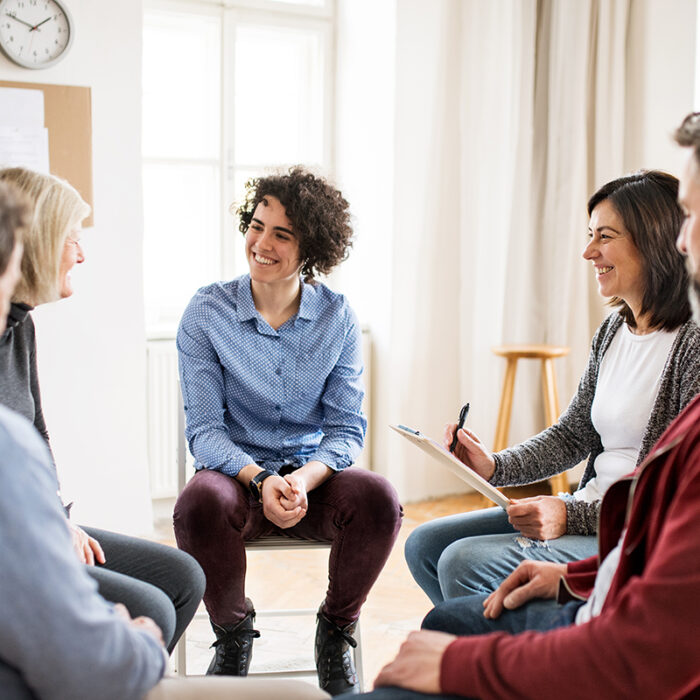 group of men and women seated in a circle and led by a facilitator who is explaining what are the benefits of group therapy