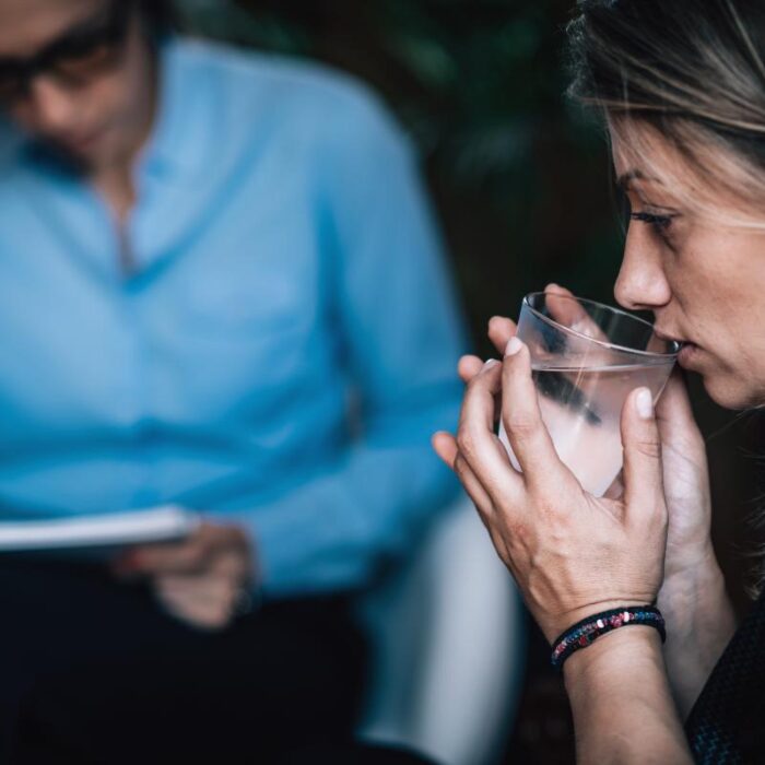 tired looking young woman taking a sip of water while therapist takes notes and explains what are the signs of prescription drug abuse